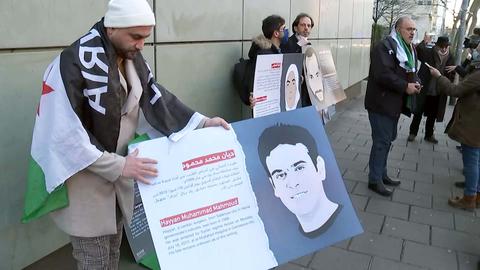 Drei Männer halten gemalte Portrait-Tafeln in die Kamera. Zudem stehen auf dem Bürgersteig daneben an eine Hauswand gelehnt weitere Portraits der Opfer von dem Anschlag in Hanau. Im Hintergund ist ein Mann mit der Presse im Gespräch.