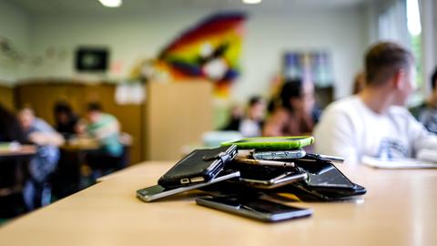 Dutzende Smartphones liegen auf einem Stapel auf einem Pult in einem Klassenraum.