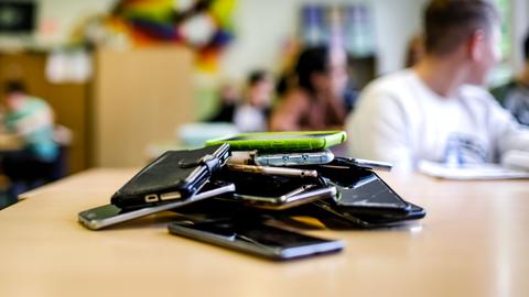 Dutzende Smartphones liegen auf einem Stapel auf einem Pult in einem Klassenraum.