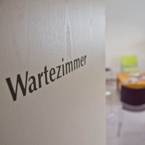 Halboffene Tür mit der Aufschrift "Wartezimmer", dahinter verschwommen leere Stühle