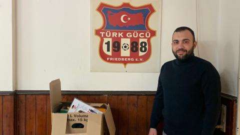 Helfer Fatih Kaplan neben Kartons mit Hilfsgütern, an der Wand das Wappen des Sportvereins Türk Gücü Friedberg e.V.