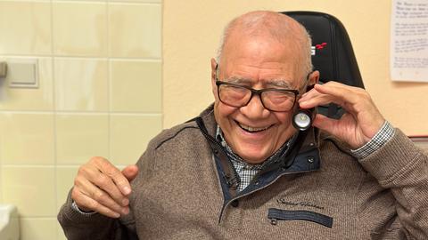 Dr. Yahia Chichakli sitzt in beiger Pullijacke in seinem Behandlungsraum, hält sich eine Taschenlampe ans Ohr und lacht