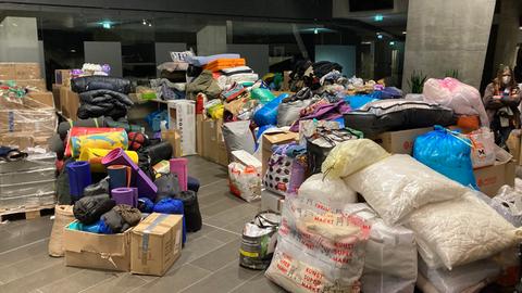 In einer Halle stehen gestapelt dutzende Kisten und Plastiktüten gefüllt mit Kissen, Isomatten, Schlafsäcken und Decken.