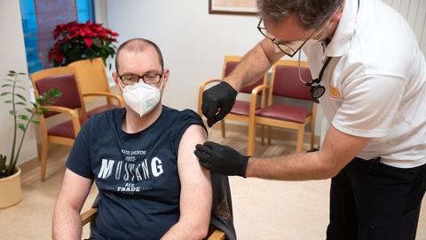 Betreuer sitzt mit Maske auf Stuhl und wird von einem Arzt geimpft
