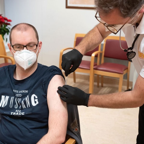 Betreuer sitzt mit Maske auf Stuhl und wird von einem Arzt geimpft