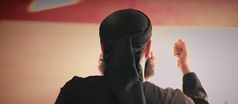 Rückenansicht eines schwarz gekleideten islamischen Predigers. Auf dem Kopf sieht man eine schwarze Kappe. Sein Bart sprießt erkennbar in alle Richtungen. Sein Gesicht ist nicht zu sehen. 