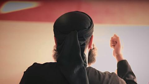 Rückenansicht eines schwarz gekleideten islamischen Predigers. Auf dem Kopf sieht man eine schwarze Kappe. Sein Bart sprießt erkennbar in alle Richtungen. Sein Gesicht ist nicht zu sehen. 