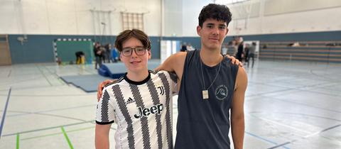 Das Bild zeigt wei Jungs mit braunen Haaren und in Sportkleidung in einer Schulsporthalle. Der Junge links ist kleiner und trägt eine Brille und ein weiß-schwarz gestreiftes Shirt, der Junge rechts trägt ein dunkelblaues Achselshirt.