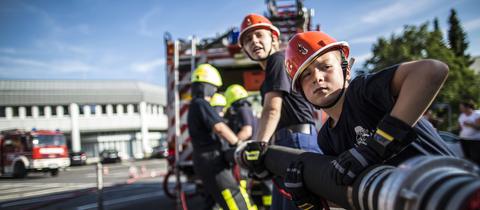 Kinder in Feuerwehrkleidung stehen an einem Löschschlauch.