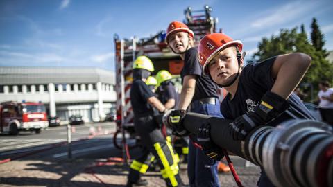 Kinder in Feuerwehrkleidung stehen an einem Löschschlauch.