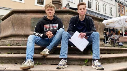 Jonathan Faust und Tom Kelwald sitzen an einem Brunnen auf der Treppe. XY hält die Klageschrift in seiner Hand. Beide tragen Jeans, Turnschuhe und dunkle Oberteile.