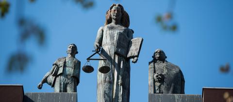 Drei Figuren auf dem Dach des Landgerichts Darmstadt. In der Mitte die blinde Justizia mit dem Richtschwert. Ihr zu beiden Seite zwei männliche Figuren. 