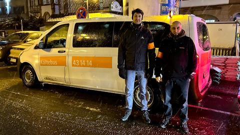 Der Kältebus-Sprinter parkt mit Warnblinker am Straßenrand in Frankfurt, Sozialarbeiter Daniel Schneider und Mourad Baadidi stehen in dicken Winterjacken vor dem Auto und lächeln in die Kamera.