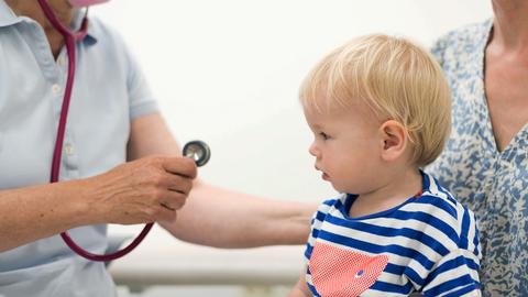 Ein Kleinkind sitzt auf dem Schoß einer Frau. Vor ihm eine Kinderärztin, die sich mit einem Stethoskop annähert.