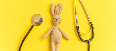 Ein Kuschel-Hase liegt auf einem gelben Untergrund. Er hat die Augen geschlossen. Neben ihm liegt ein Stethoskop.