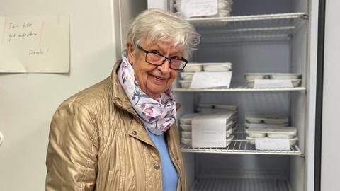 Eine ältere Frau steht vor einem offenen Kühlschrank, darin liegen viele Essensportionen in Einwegverpackungen. Sie schaut in die Kamera.