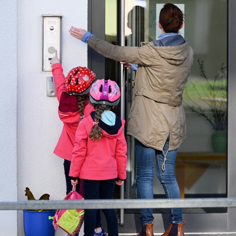 Mutter mit Kindern stehen vor und klingeln an einer Kita-Tür.