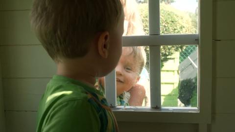 Einer der Zwillinge steht in einem Holzhäuschen, der andere schaut durch das Fenster nach drinnen.