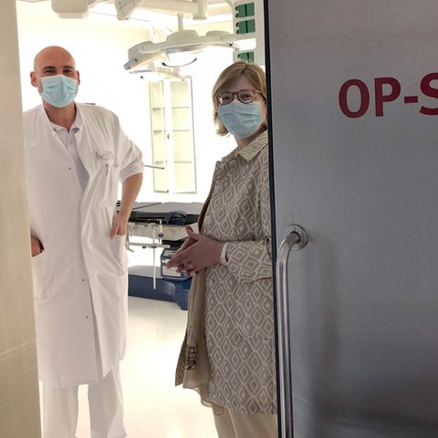 Ein Arzt und eine Businessfrau mit Masken stehen im Rahmen einer Tür, die zum OP führt, und lächeln in die Kamera. Auf der Tür steht "OP-SAAL 6". Der OP ist sehr hell erleuchtet.
