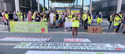 Streikende in gelben Westen mit Transparenten wie "Wenn wir streiken, steht die Welt still" und "Mehr von uns ist besser für alle".