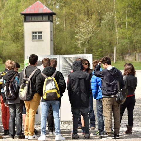 Eine Gruppe von Jugendlichen steht auf einem Platz in einem ehemaligen Konzentrationslager, im Hintergrund ein Wachturm