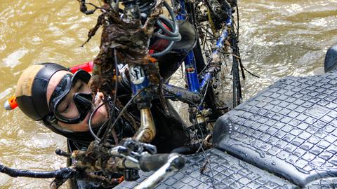 Eine Frau in Tauchkleidung, deren Oberkörper aus braunem Wasser ragt. In der Hand hält sie Schrott, Fahrradteile sind zu erkennen.