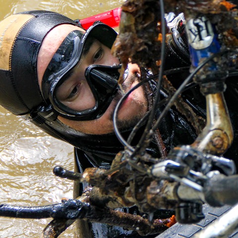 Eine Frau in Tauchkleidung, deren Oberkörper aus braunem Wasser ragt. In der Hand hält sie Schrott, Fahrradteile sind zu erkennen.