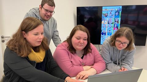 Drei Frauen sitzen an einem Tisch vor einem Laptop und schauen auf den Bildschirm. Hinter ihnen steht ein Mann und schaut ebenfalls auf den Computer.