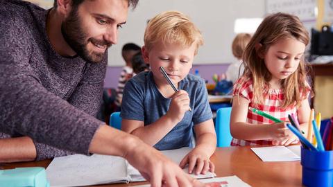 Ein Lehrer sitzt neben einer Schülerin und einem Schüler an einem Tisch und erklärt.