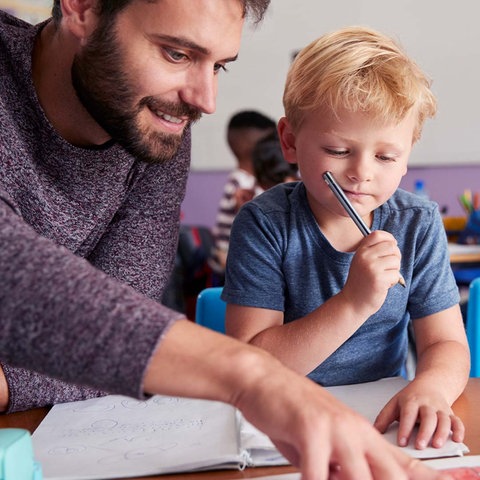 Ein Lehrer sitzt neben einer Schülerin und einem Schüler an einem Tisch und erklärt.