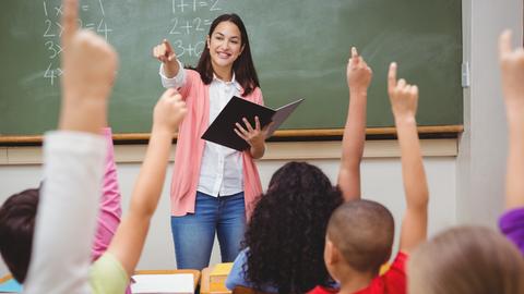 Eine Lehrerin steht im Klassenraum vor Schülerinnen und Schüler und zeigt auf etwas.