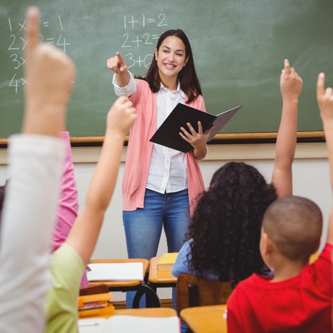 Eine Lehrerin steht im Klassenraum vor Schülerinnen und Schüler und zeigt auf etwas.