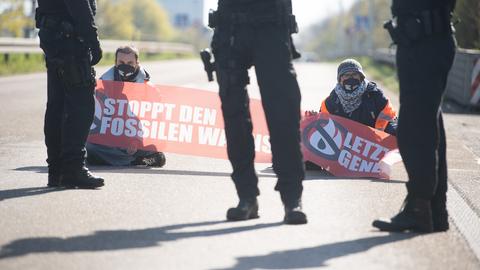 Zwei Menschen sitzen auf der Fahrbahn der Autobahn und halten ein Transparent mit der Aufschrift "Stoppt den fossilen Wahnsinn" in der Hand. Davor stehen drei Polizisten.