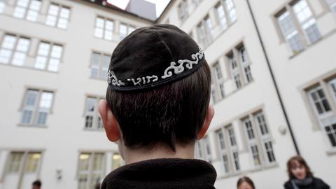 Foto eines männlichen Hinterkopfes, der eine Kippa trägt. Im Hintergrund unscharf ein historisches Schulgebäude mit großen Fenstern.