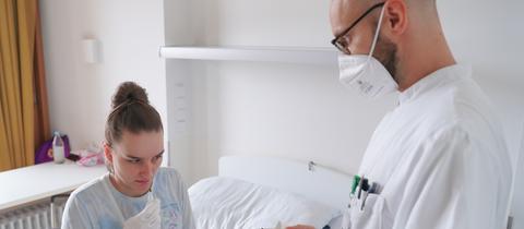 Eine Patientin sitzt auf einem Krankenhausbett, neben ihr ein Arzt in weißem Kittel.