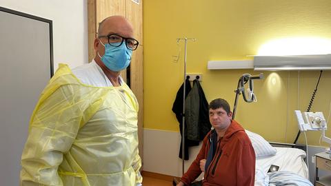 Ein Arzt steht mit Maske und Kittel neben einem Patienten, der auf dem Bett sitzt
