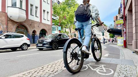 Junge Radfahrerin mit einem Rucksack, von hinten fotografiert, die auf einem Radweg in einer Geschäftsstraße fährt. 