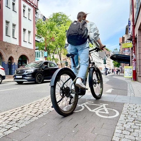 Junge Radfahrerin mit einem Rucksack, von hinten fotografiert, die auf einem Radweg in einer Geschäftsstraße fährt. 