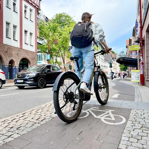 Junge Radfahrerin mit einem Rucksack, von hinten fotografiert, die auf einem Radweg in einer Geschäftsstraße fährt. 