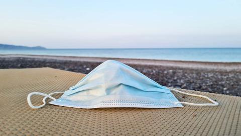 Eine OP-Maske liegt auf einer Liege an einem Strand mit Meer und Horizont im Hintergrund.
