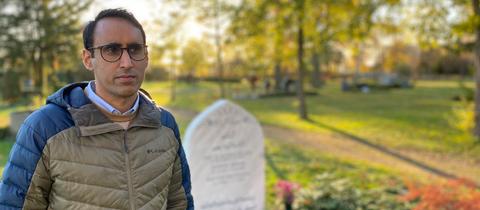 Ein Mann steht auf einem Friedhof neben einem weißen steinernen Grabstein und schaut in die Kamera.