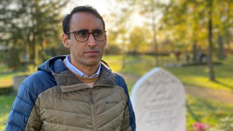 Ein Mann steht auf einem Friedhof neben einem weißen steinernen Grabstein und schaut in die Kamera.