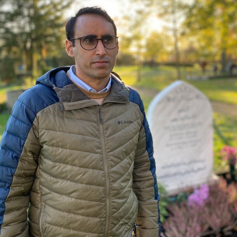 Ein Mann steht auf einem Friedhof neben einem weißen steinernen Grabstein und schaut in die Kamera.