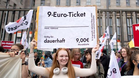 Eine junge Frau hält ein überdimensionales 9-Euro-Ticket auf einer Demo hoch und lacht.