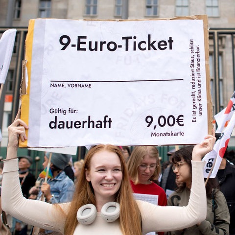 Eine junge Frau hält ein überdimensionales 9-Euro-Ticket auf einer Demo hoch und lacht.