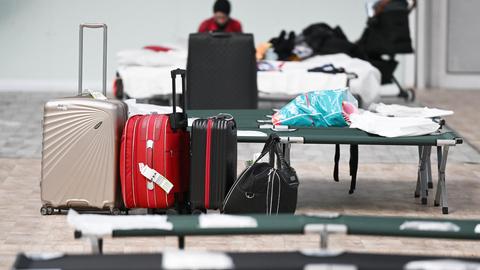 Im Bildvordergrund: Pritschen und Koffer in einer Notunterkunft in Frankfurt. Unscharf um Bildhintergund der Raum mit einem Menschen, der auf einem Notbett sitzt.