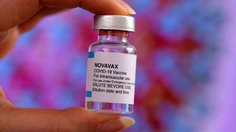 Eine Hand hält ein mit klarer Flüssigkeit gefülltes Fläschchen mit der Aufschrift "Novavax Covid-19 Vaccine"