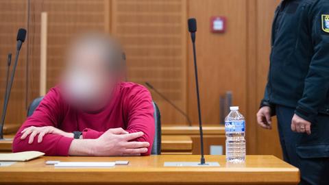 Ein Mann in rotem Pullover sitzt in einem Gerichtssal, neben ihm steht ein Justizbeamter in Uniform.