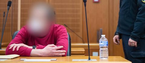 Ein Mann in rotem Pullover sitzt in einem Gerichtssal am Tisch, neben ihm steht ein Justizbeamter in Uniform.