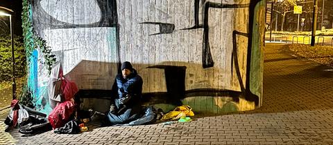 Der obdachlose Christian Endres sitzt in dicker Winterjacke, mit Mütze und Schlafsack unter einer Brücke, um sich vor Schneeregen zu schützen. 